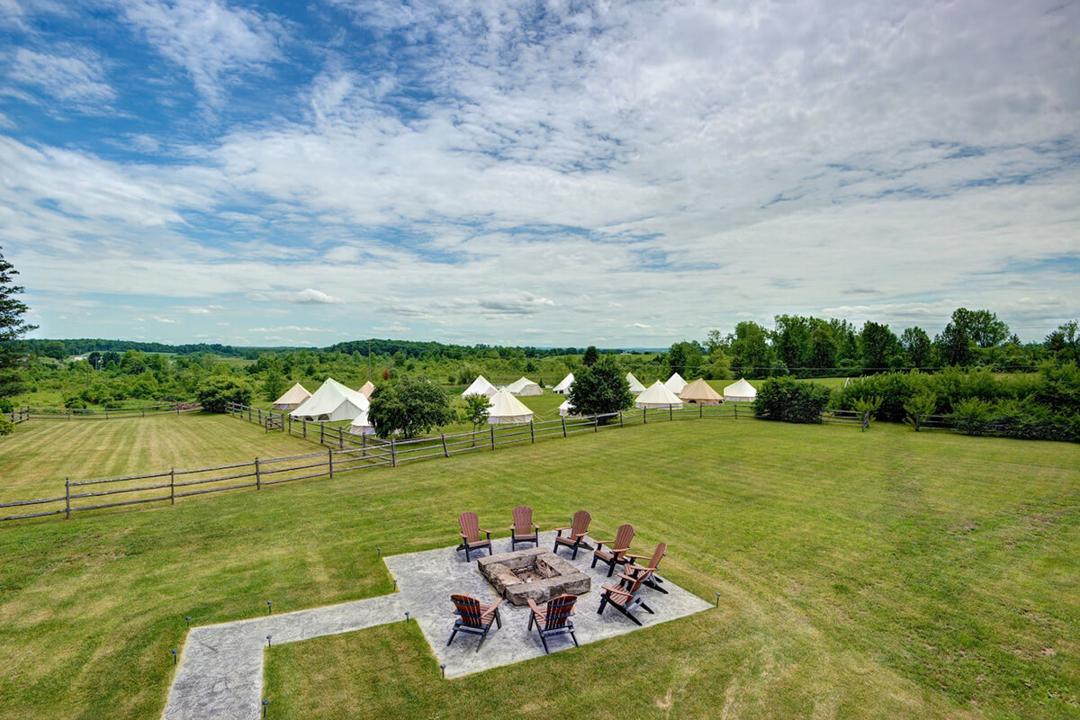 Aerial view of fire pit with chairs in green pasture with scenic mountain range in background.